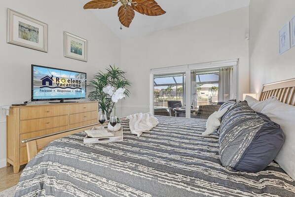 A hotel room with a bed, a wooden dresser, a television displaying a sign, a potted plant, and a view of the outdoors through a sliding door.