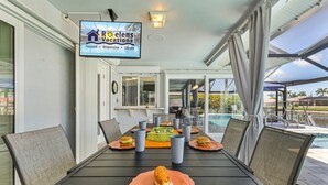 Un coin repas avec une table dressée pour deux personnes, un écran de télévision affichant un logo, et une vue sur une piscine et des palmiers.
