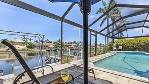 Un espace aménagé au bord de la piscine, avec un patio fermé par des baies vitrées, une table en verre sur laquelle se trouve une boisson, et une vue sur une étendue d’eau et des bâtiments en arrière-plan.