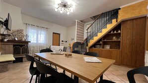 Een eethoek met een houten tafel, zwarte stoelen en een kroonluchter. Er is een trap met een houten leuning die naar een lager gelegen niveau leidt.