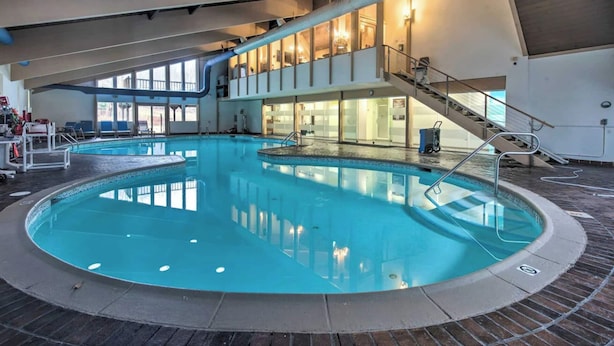 Piscina interna com água cristalina, escada que leva a um segundo nível e vista para o exterior por grandes janelas.