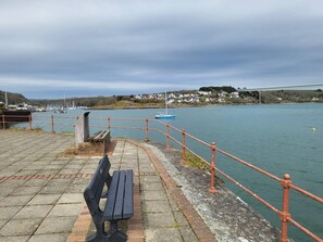 A waterfront promenade with a bench, a railing, and a view of boats and houses across the water.