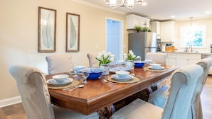Uma sala de jantar com uma mesa de madeira posta para quatro pessoas, cercada por cadeiras claras. Há quadros com impressões botânicas na parede, um lustre sobre a mesa e uma vista para a cozinha adjacente.