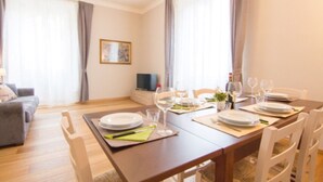 A dining area with a wooden table set for four, surrounded by white chairs. There is a television on a stand against the wall, and a sofa is visible in the background. - Cittadella Apartment (Florence)