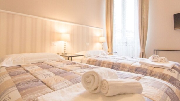 A hotel room with two beds, each with a rolled towel, a nightstand with a lamp, and a window with curtains. - Cittadella Apartment (Florence)