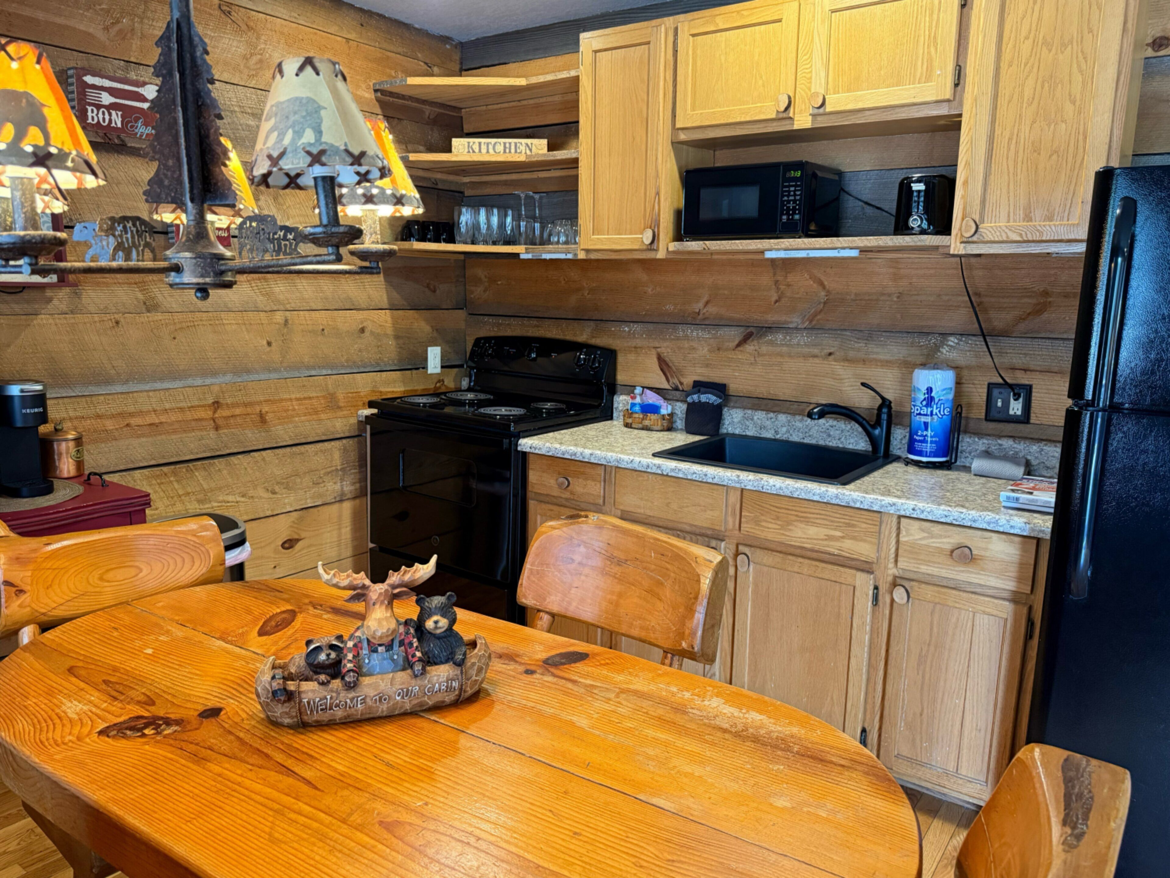 A wooden kitchen with a stove, microwave, and sink. There is a wooden table with a bear figurine.