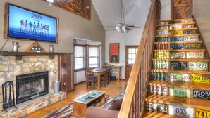 A spacious living room with a stone fireplace, a television mounted above it, and a collection of license plates on a wooden staircase.