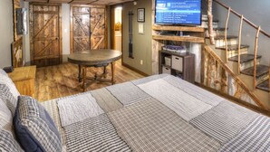 A rustic room with a wooden floor, a round table, a bed with striped bedding, and a TV on a wooden shelf.