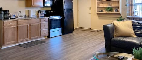 Una cocina moderna con armarios de madera, un refrigerador negro y un microondas. Hay una mesa de centro de vidrio, una alfombra y un sofá con un cojín.