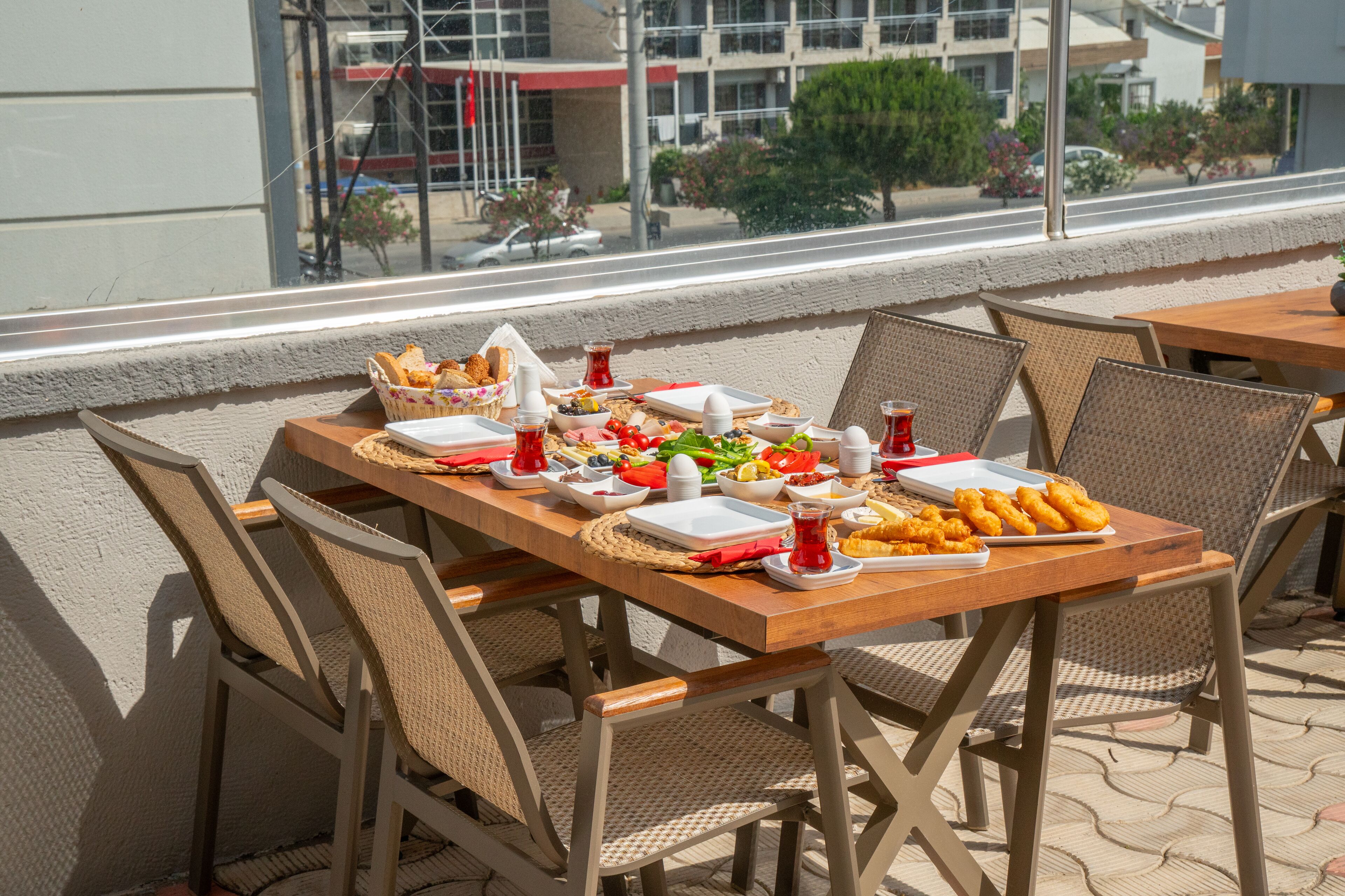 Una mesa de madera con platos, tazones y vasos, rodeada de sillas de mimbre, en un patio exterior con vistas a edificios y árboles.