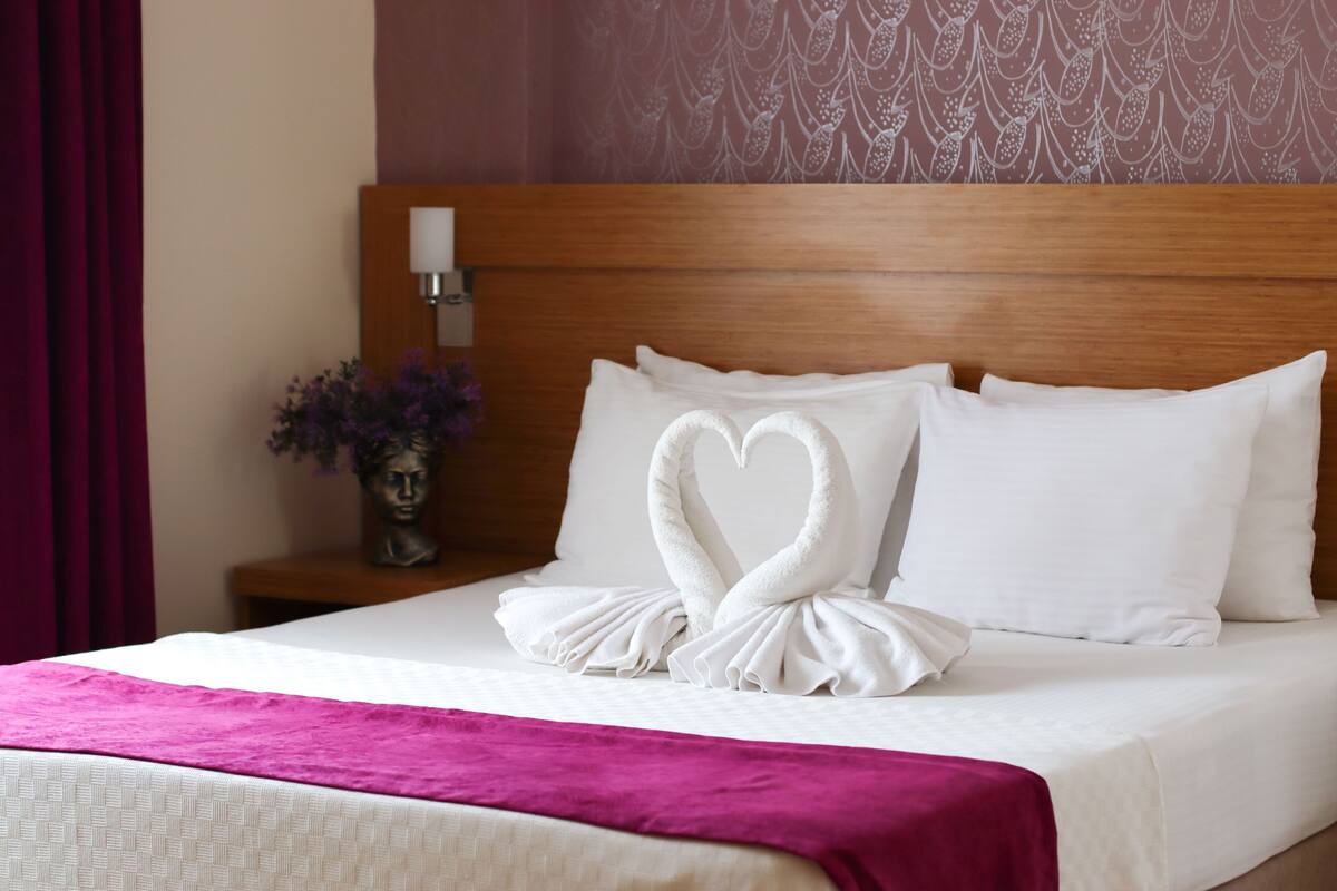 Una habitación de hotel con una cama bien hecha, un cabecero de madera, una mesita de noche con una lámpara y un jarrón con flores.