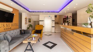 A hotel reception area with a sofa, armchair, reception desk, and a television mounted on the wall. - Siriusmi hotel (Cesme)