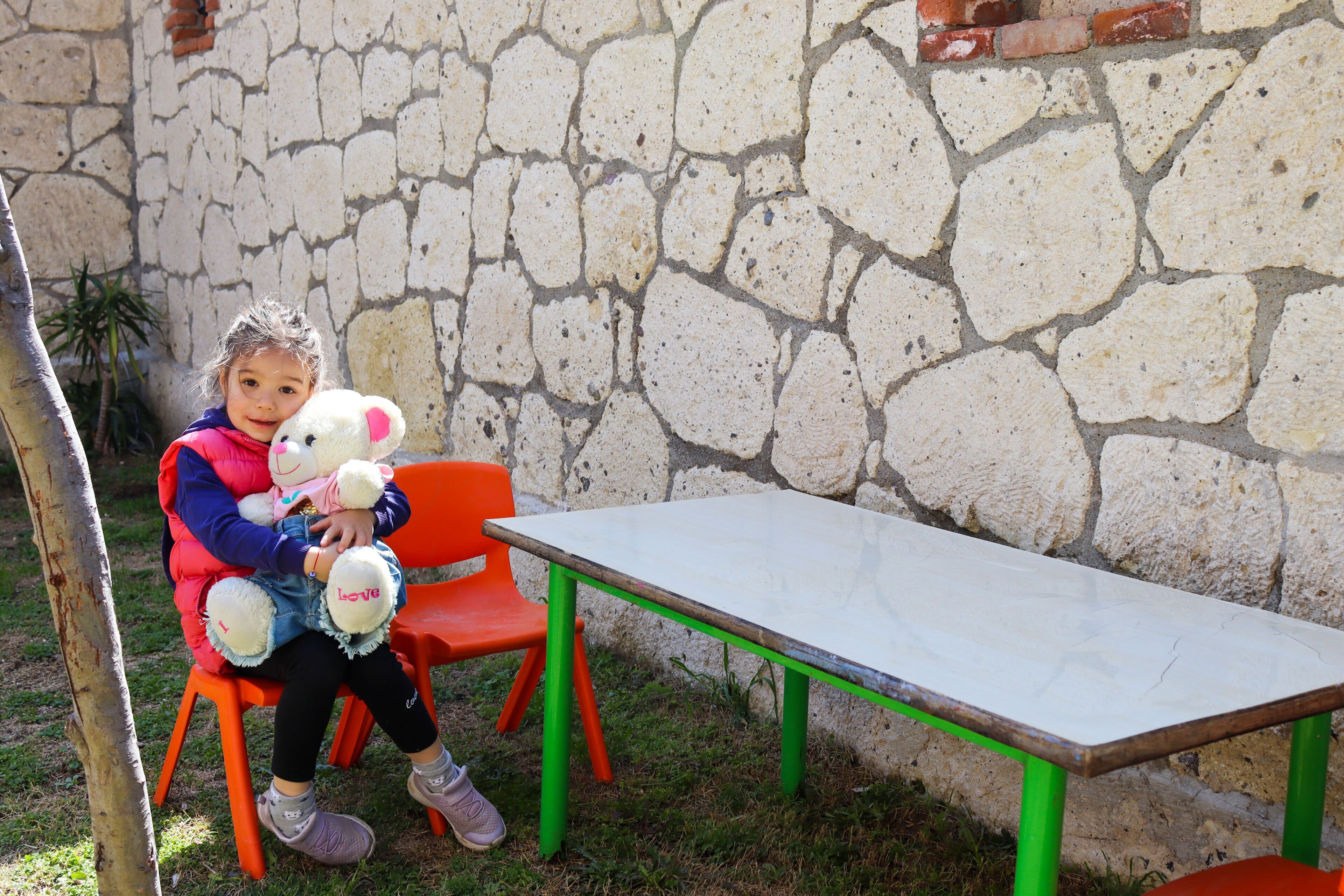 A child sitting on an orange chair with a teddy bear, next to a white table with green legs, in front of a stone wall.