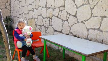A child sitting on an orange chair with a teddy bear, next to a white table with green legs, in front of a stone wall.