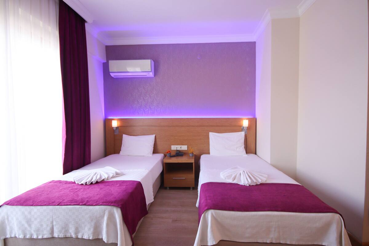 Habitación de hotel con dos camas, cortinas moradas, cabecero de madera y un equipo de aire acondicionado.
