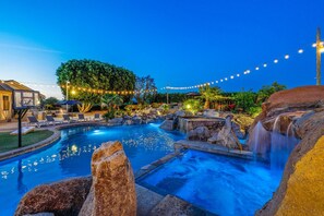 Une piscine avec une cascade, entourée de rochers et d’arbres, et des guirlandes lumineuses en arrière-plan.