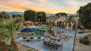 A pool area with lounge chairs, a dining table, and a thatched-roof structure. - The Bentley Estate: Festivals, Karaoke, Grotto, Sauna, Putting Green, BBQ! (Bermuda Dunes)