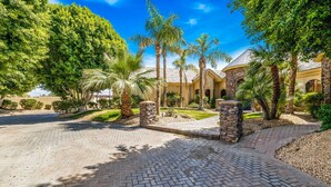 A paved driveway leading to a building with stone pillars and palm trees. - The Bentley Estate: Festivals, Karaoke, Grotto, Sauna, Putting Green, BBQ! (Bermuda Dunes)