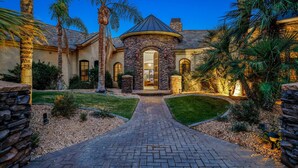A well-lit, stone-clad house with a curved entrance, surrounded by a landscaped garden and palm trees. - The Bentley Estate: Festivals, Karaoke, Grotto, Sauna, Putting Green, BBQ! (Bermuda Dunes)