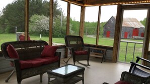 Un patio cubierto con mobiliario de mimbre, una mesa de vidrio y vistas a un granero y follaje.