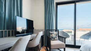 Habitación de hotel con un ventanal grande, un escritorio con televisor, una silla, una cama y un balcón con mobiliario exterior.