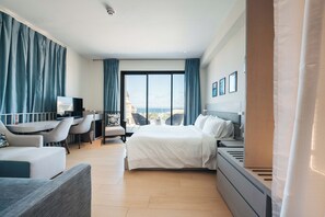 A modern hotel room with a large bed, a desk, and a seating area with chairs. There is a view of the ocean through large windows. - Elements Hotel & Shops (Willemstad)