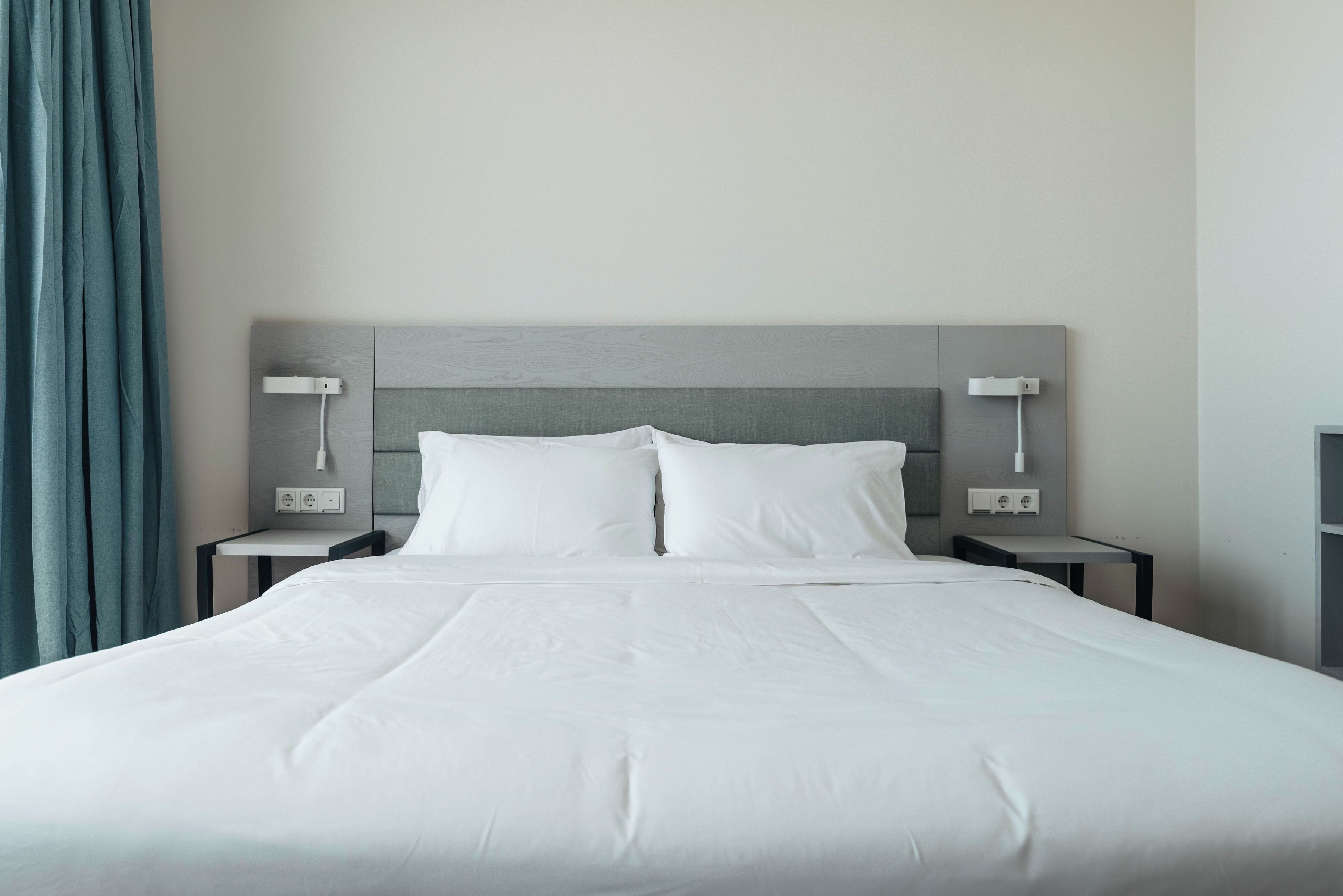 Una cama bien hecha con sábanas blancas, dos mesitas de noche con lámparas y un cabecero gris.