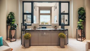 Un área de recepción moderna con un mostrador central, dos ventanas amplias y plantas en macetas.