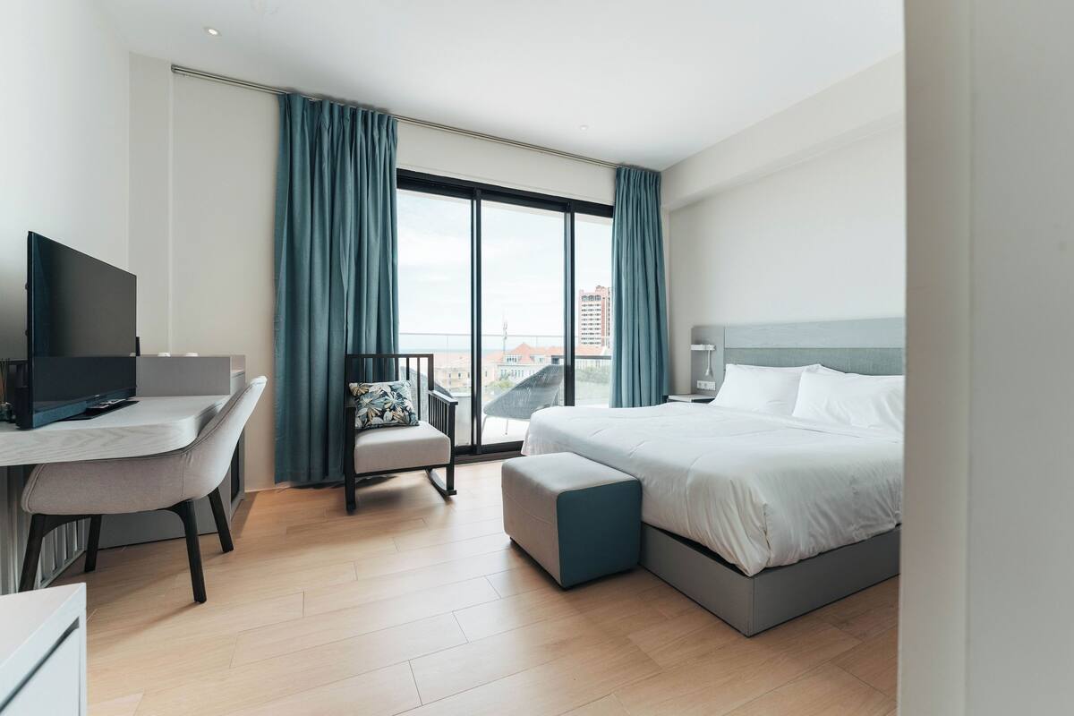 A modern hotel room with a bed, a desk, a chair, and a large window with curtains.