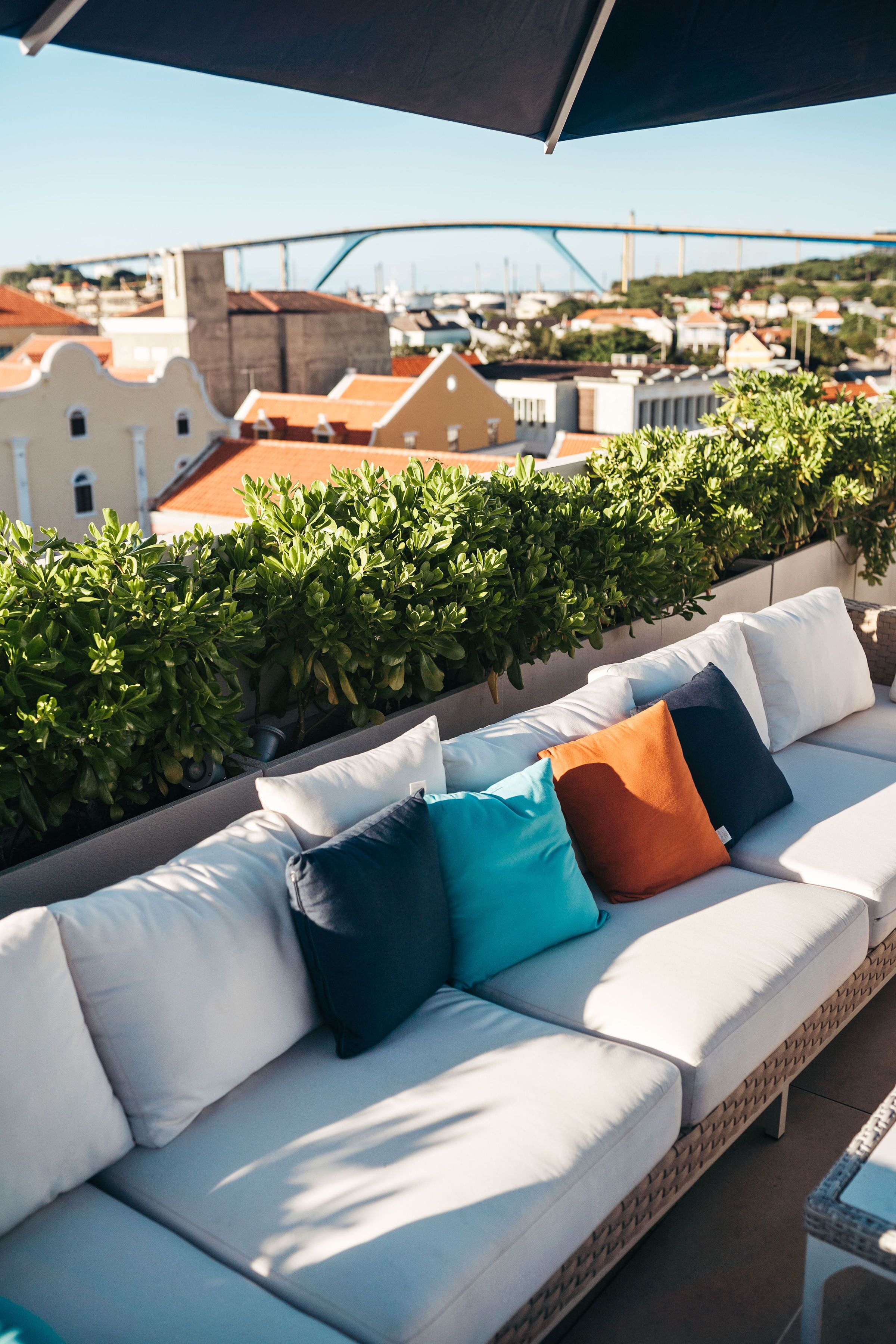 Un sofá blanco para exteriores con cojines coloridos, una mesa de vidrio y una vista de edificios y un puente al fondo.