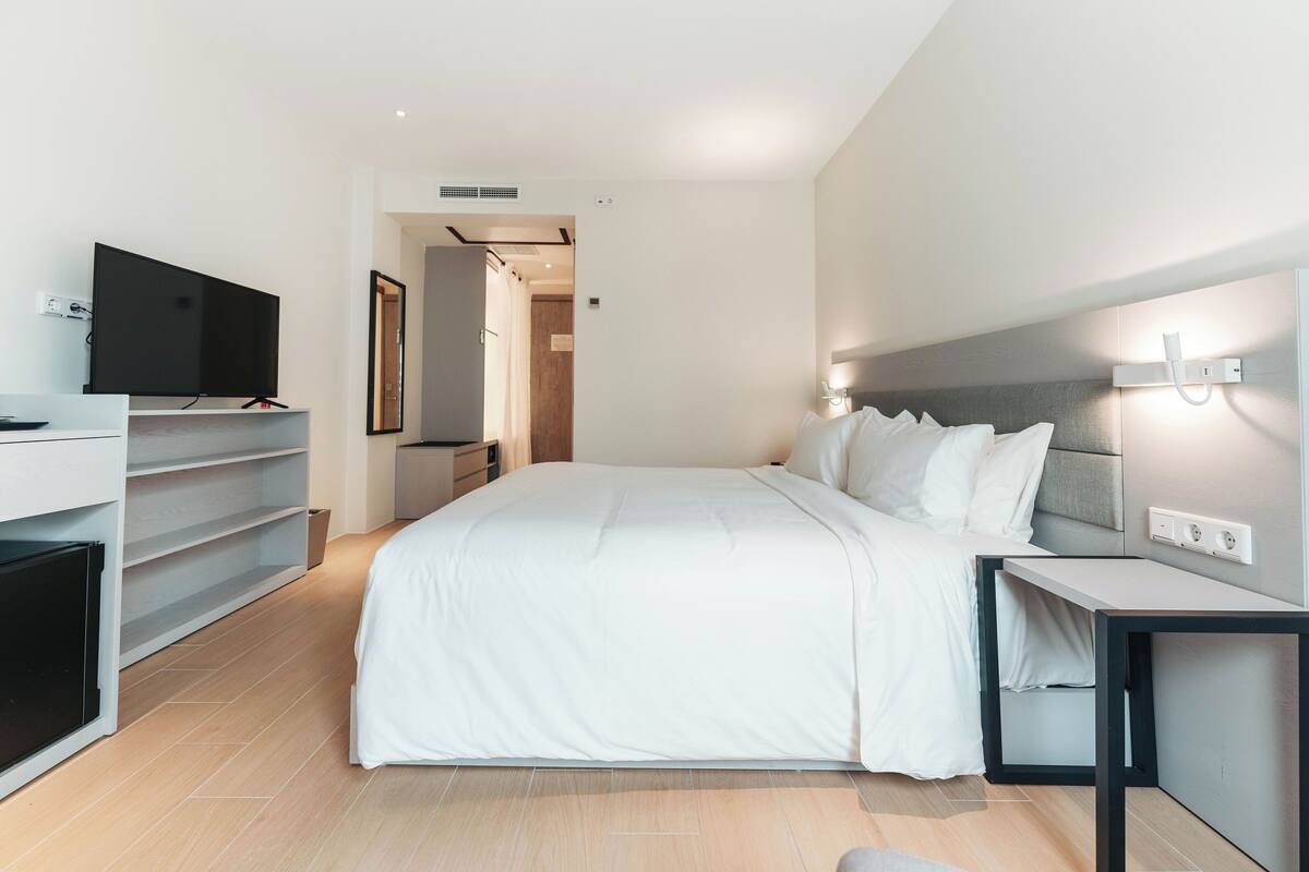 A modern hotel room with a large bed, a flat-screen TV, a bedside table, and a built-in desk.