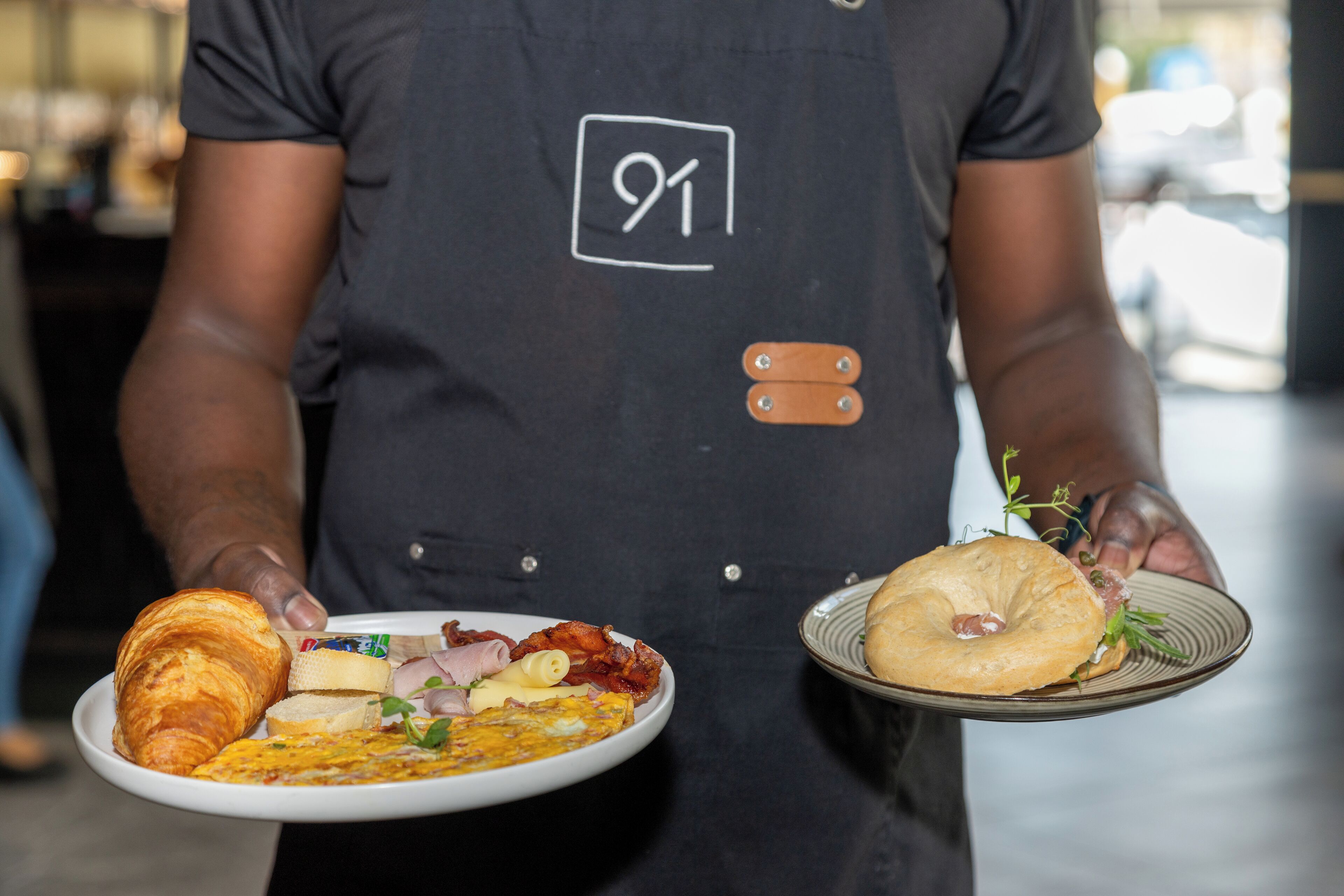 A person holding two plates of food, one with a croissant and the other with a bagel and a sandwich.