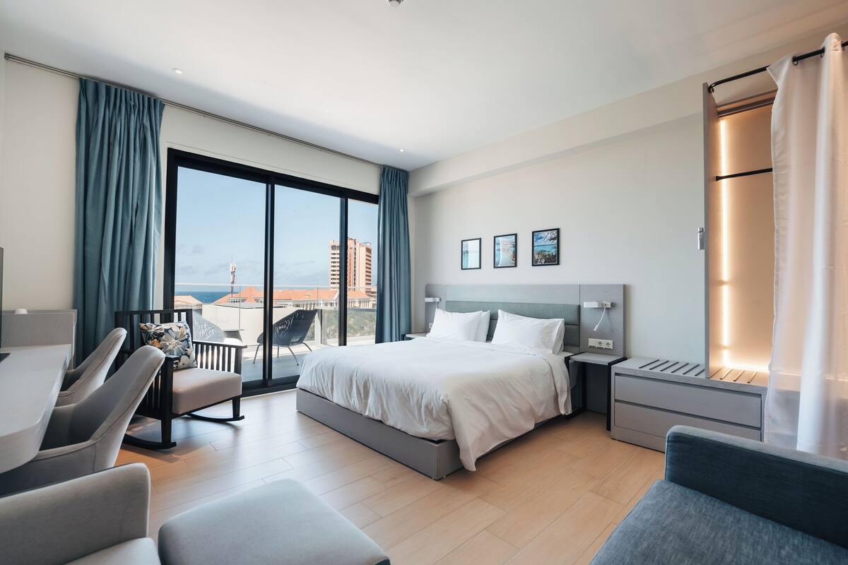 A modern hotel room with a large bed, a desk, a chair, and a view of the cityscape.