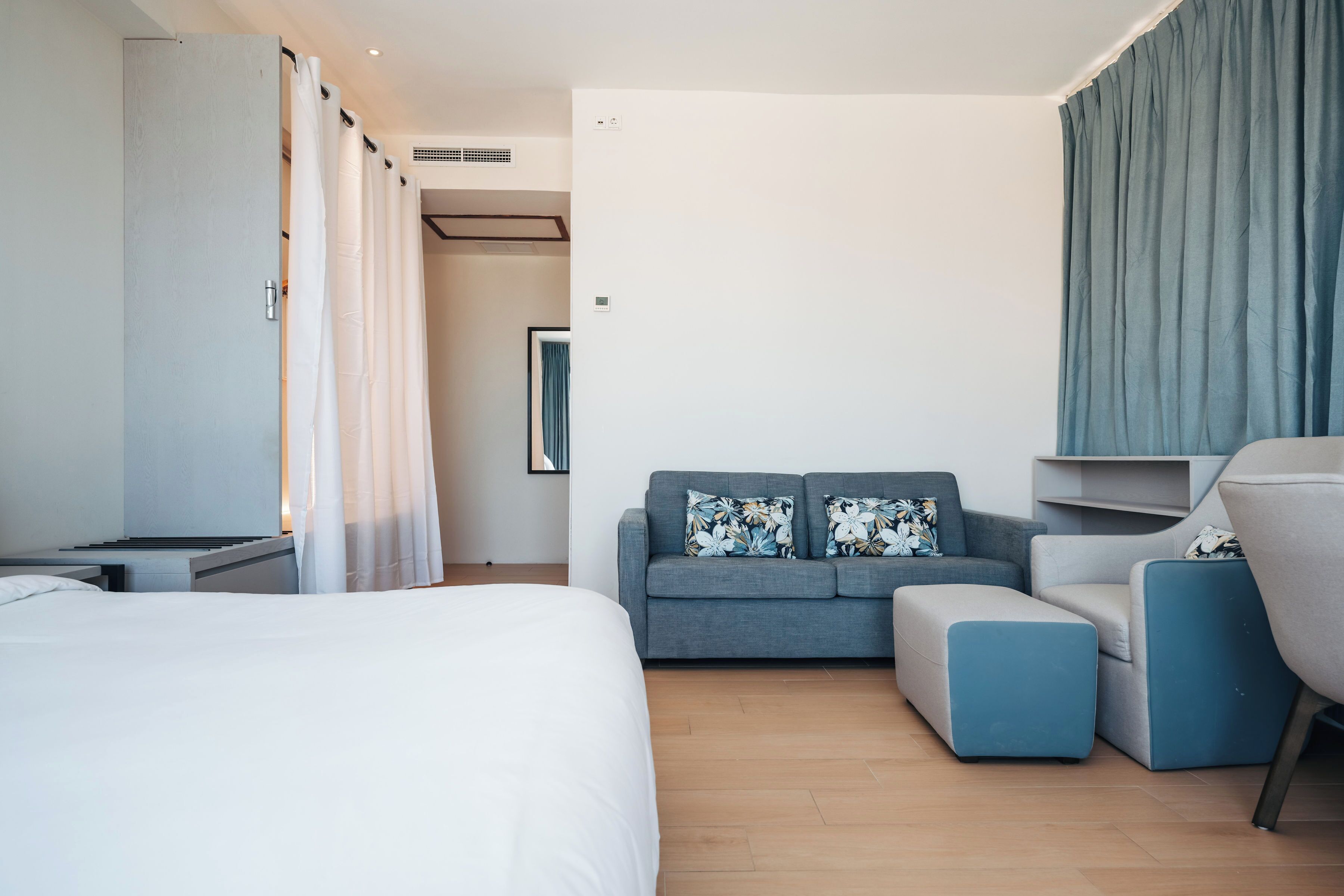 Una camera d'albergo moderna con un letto grande, un divano grigio con cuscini floreali, una poltrona blu e un'ampia finestra con tende.