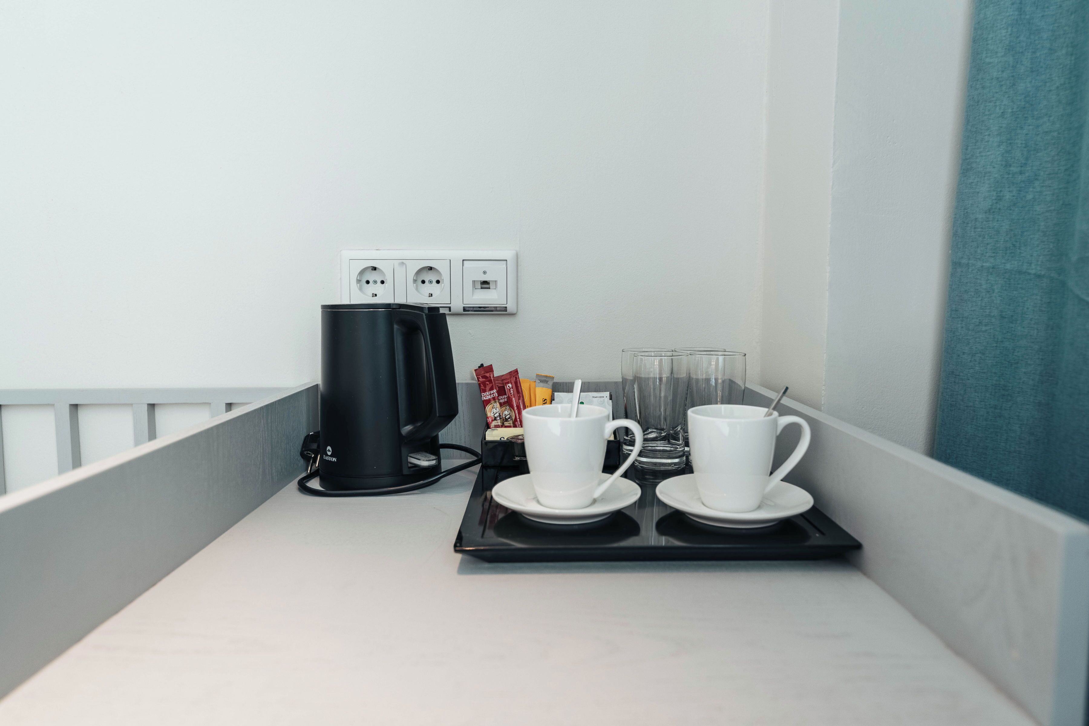 A hotel room with a kitchenette area featuring a coffee maker, two white cups with saucers, a glass of water, and a tray with a coffee pot and a packet of sugar.