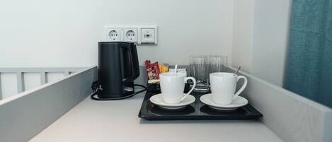 A hotel room with a kitchenette area featuring a coffee maker, two white cups with saucers, a glass of water, and a tray with a coffee pot and a packet of sugar.