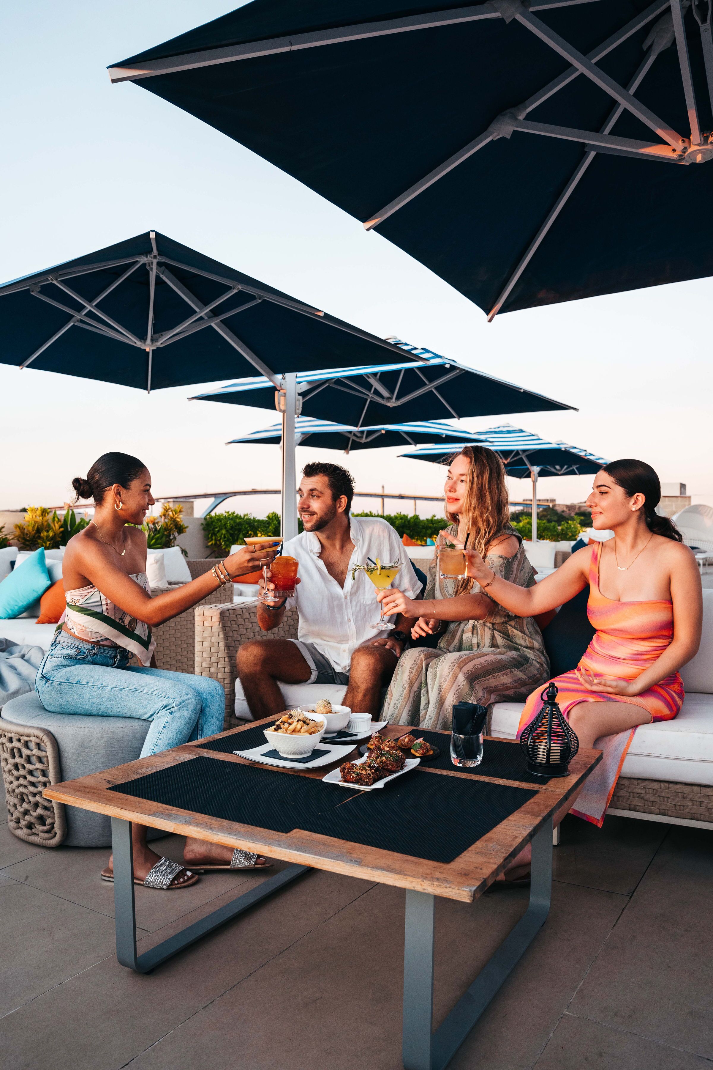 A group of people sitting at an outdoor table with umbrellas, enjoying drinks and food.