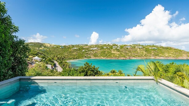 Une piscine à l’eau d’un bleu cristallin, entourée d’une végétation luxuriante, offrant une vue sur une plage turquoise et une colline parsemée de maisons.