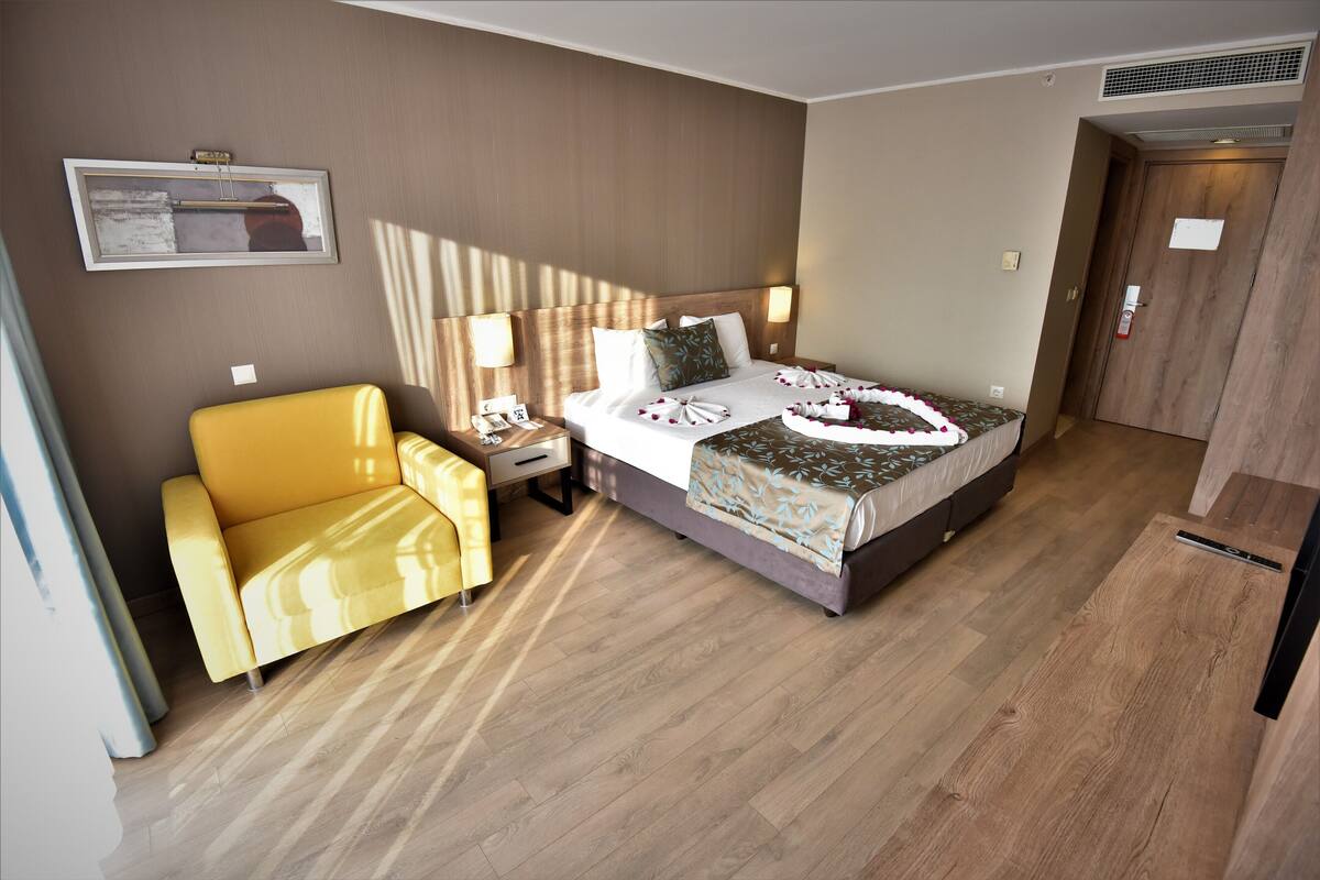 Ein Hotelzimmer mit Bett, gelbem Sessel, Nachttisch und Fenster mit Jalousien.