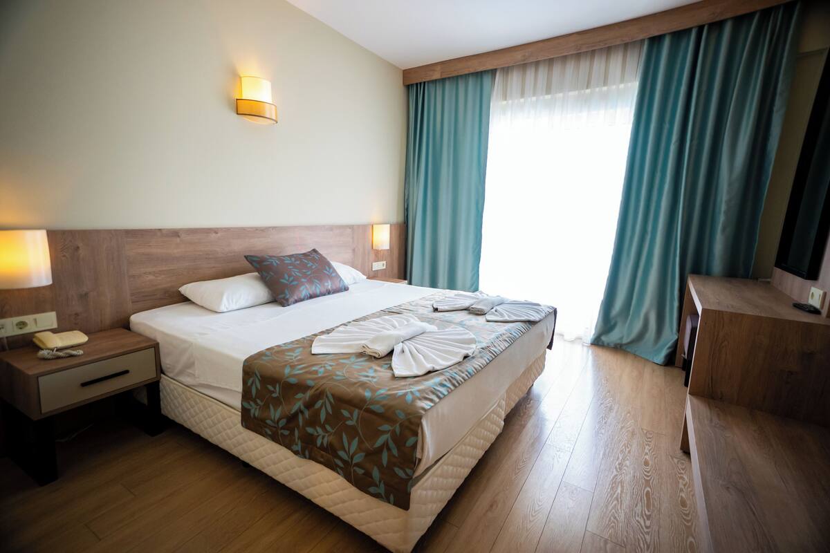 Ein Hotelzimmer mit Bett, Nachttischlampen, einem Holzkopfteil, einem Schreibtisch und einem fenster mit Vorhängen.