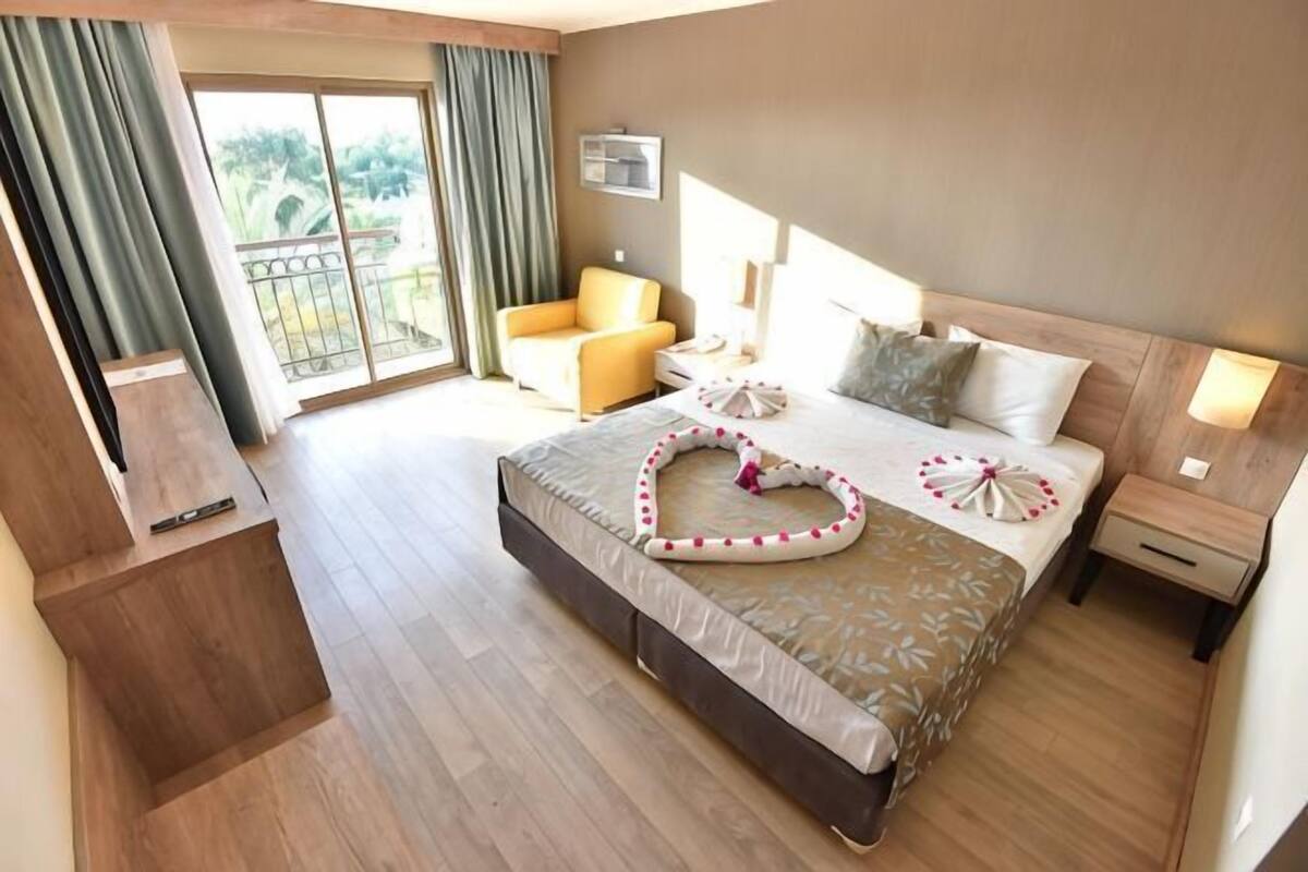 Ein Hotelzimmer mit Bett, Sessel, Nachttisch, Fernseher und einem Balkon mit Vorhängen.