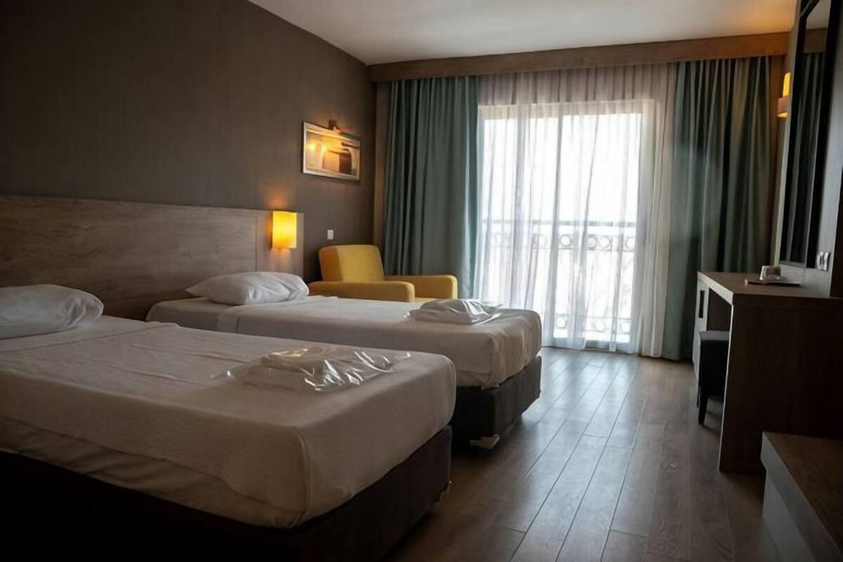 Ein Hotelzimmer mit zwei Betten, einem Sessel, einem Schreibtisch und einem großen Fenster mit Vorhängen.