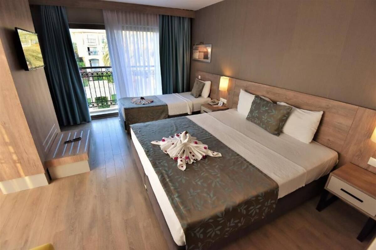 Ein Hotelzimmer mit zwei Betten, einem Fernseher, einem Balkon mit Vorhängen und einem Holzboden.