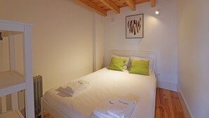 Een net opgemaakt bed met witte lakens en groene kussens, een witte radiator en een houten plafond.