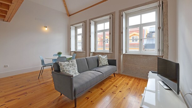 Een moderne woonkamer met een grijze bank, houten vloer en grote ramen.