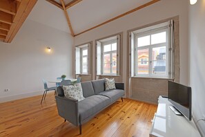 A modern living room with a grey sofa, wooden flooring, and large windows.