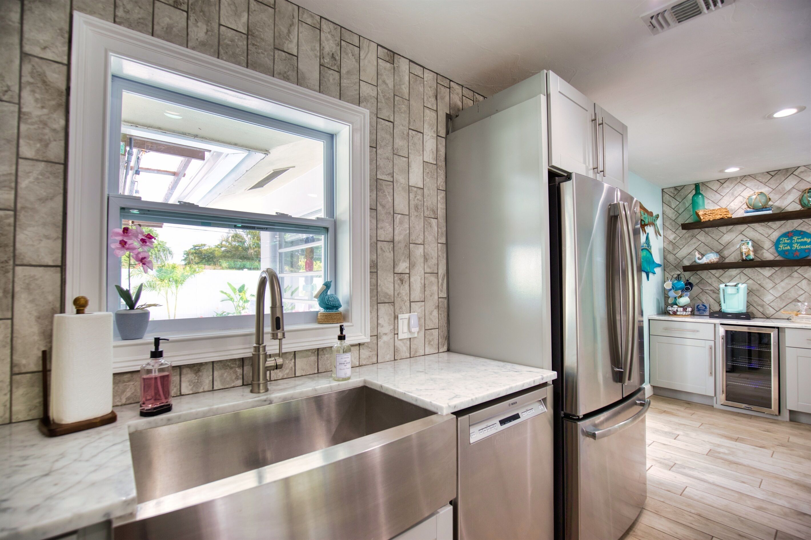 A modern kitchen with a stainless steel refrigerator, a sink, and a window with a view of greenery.