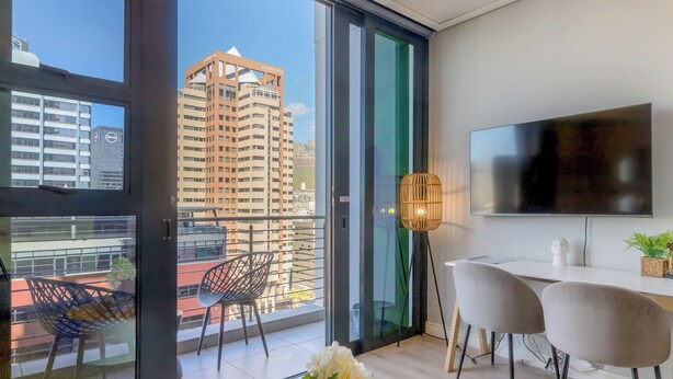 Un salon moderne doté d’un grand téléviseur, de deux fauteuils, d’une table à manger et d’un balcon offrant une vue sur la ville.