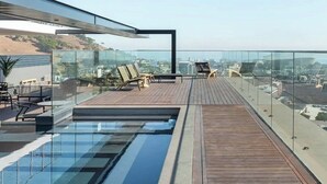 Une piscine sur le toit, dotée d’une terrasse en bois et de chaises longues, le tout entouré de rampes de protection en verre.