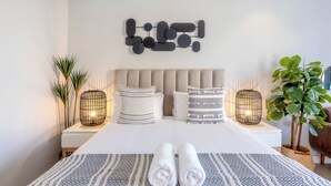 Un lit bien fait, agrémenté de serviettes pliées, une œuvre d’art moderne et abstraite accrochée au mur, et deux tables de chevet ornées de lampes et de plantes.